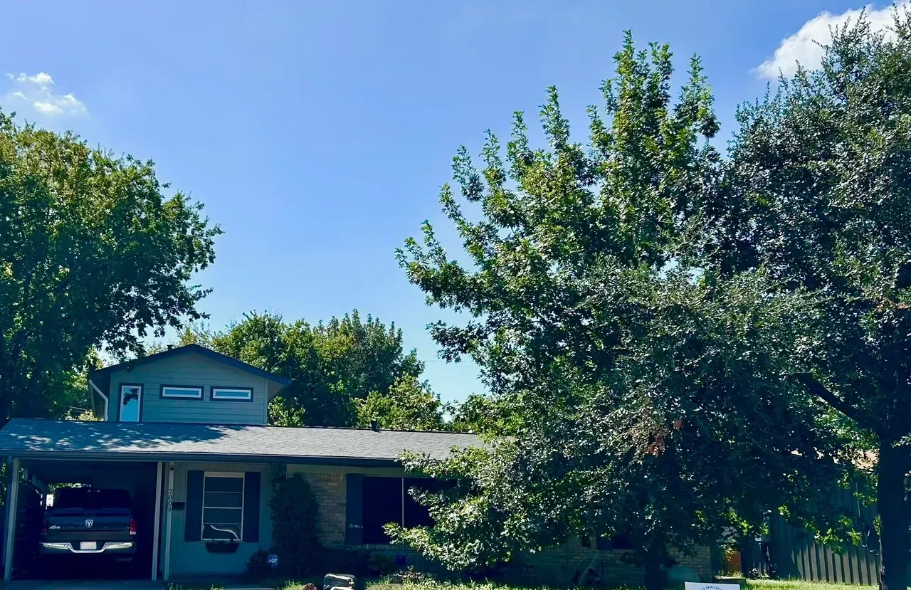 House with a carport, trees, and a blue sky. A truck is parked in the carport.