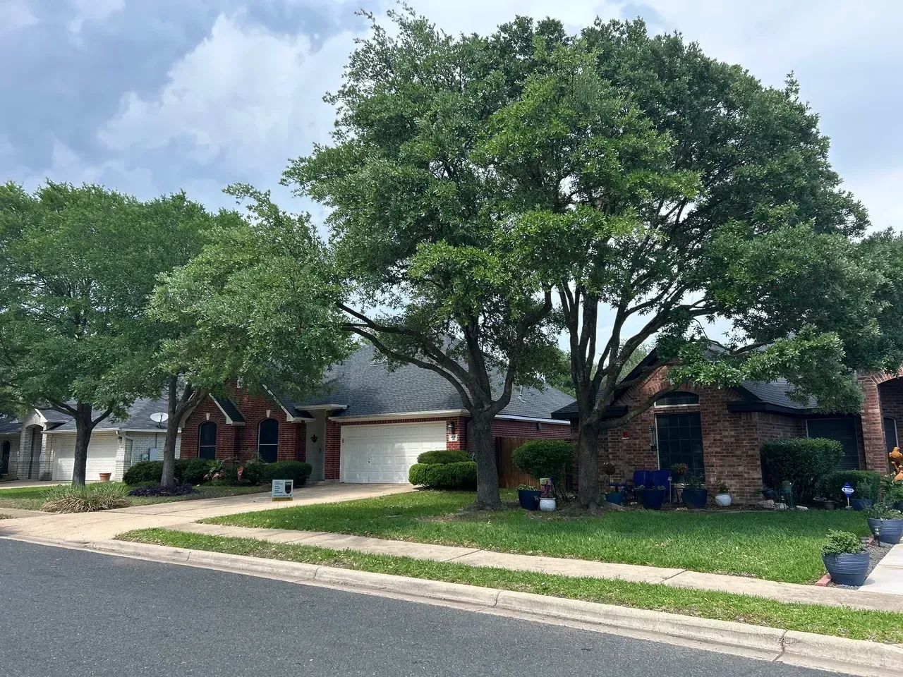 Houses with brick exteriors, lush green lawns, and mature trees line a residential street under a cloudy sky.