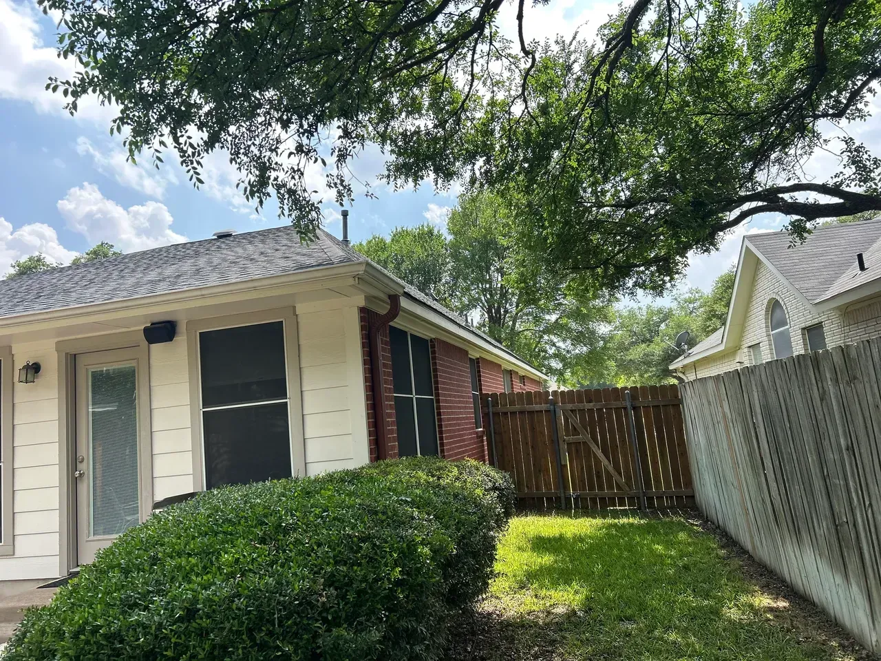 Exterior of a house with red brick and cream siding, next to a wooden fence and yard, under tree branches.