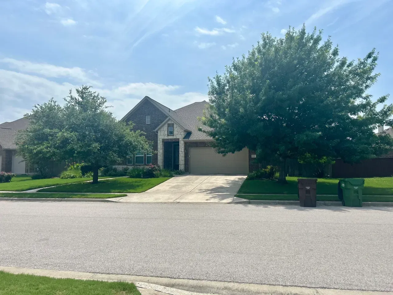 Suburban house with a driveway, lawn, and trees under a blue sky. Two trash bins in the front.