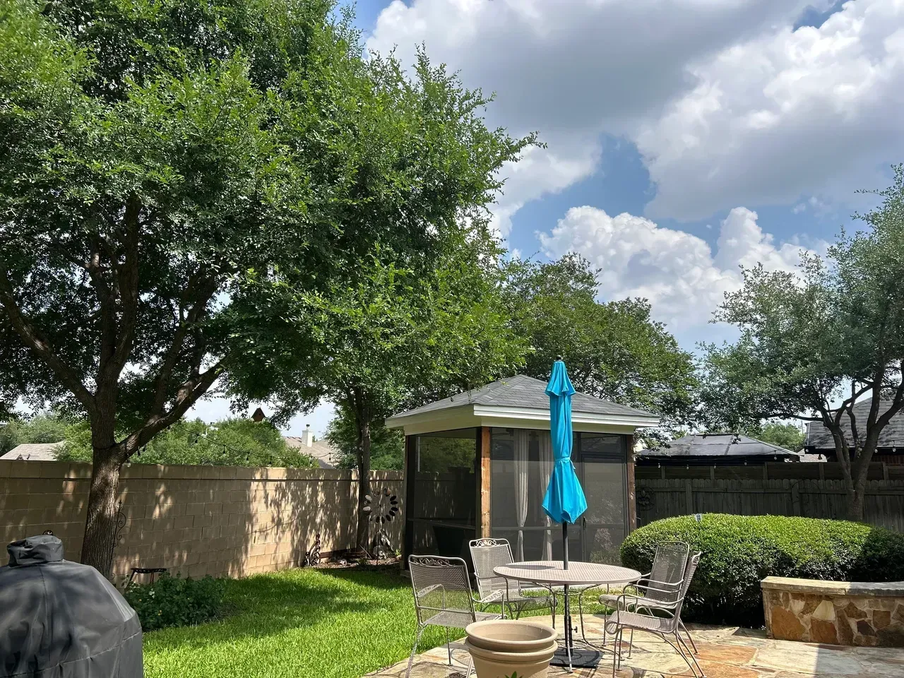 Backyard scene: gazebo with blue umbrella, patio furniture, green grass, trees, cloudy sky.