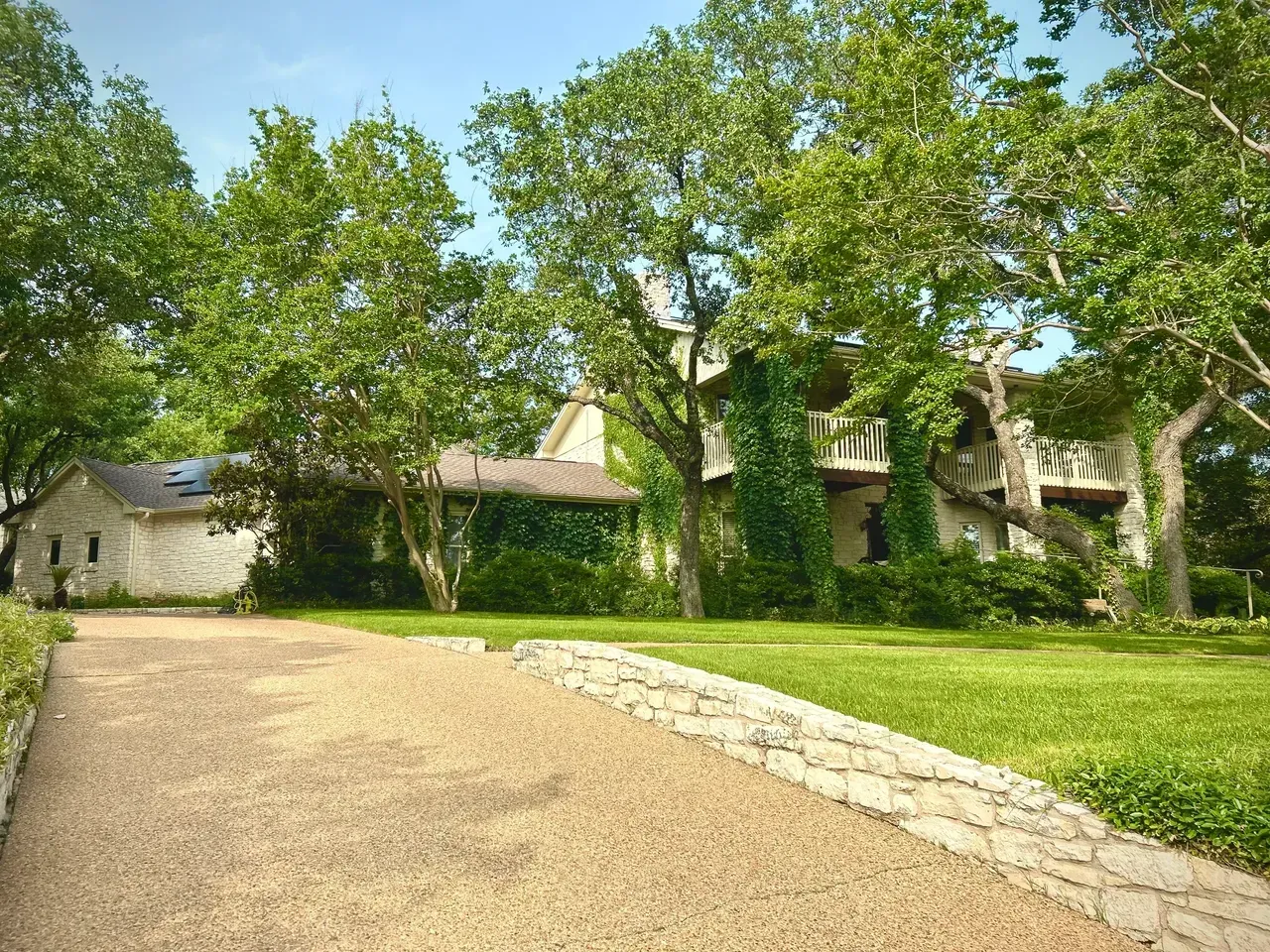 Gravel driveway leading to a two-story house with a stone exterior covered in greenery. Lush green trees surround the home.
