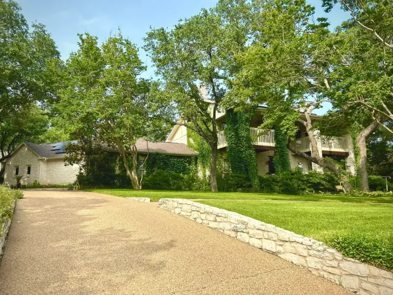 Gravel driveway leading to a stone house surrounded by green trees and grass on a sunny day.