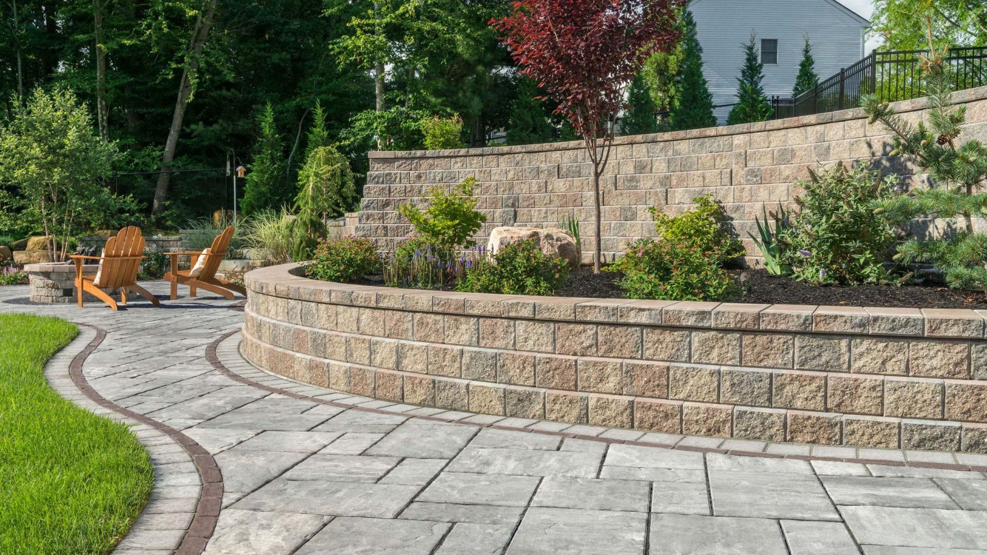 A curved stone retaining wall borders a patio with wooden chairs, surrounded by landscaping and trees on a sunny day.