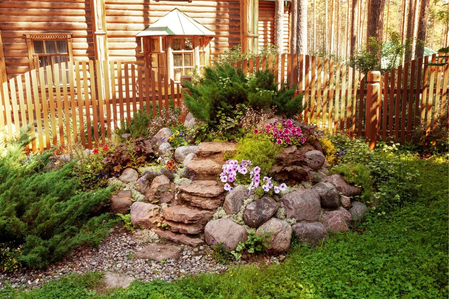 A rock garden feature with small stone steps, colorful flowers, and evergreens in front of a wooden fence and log house.