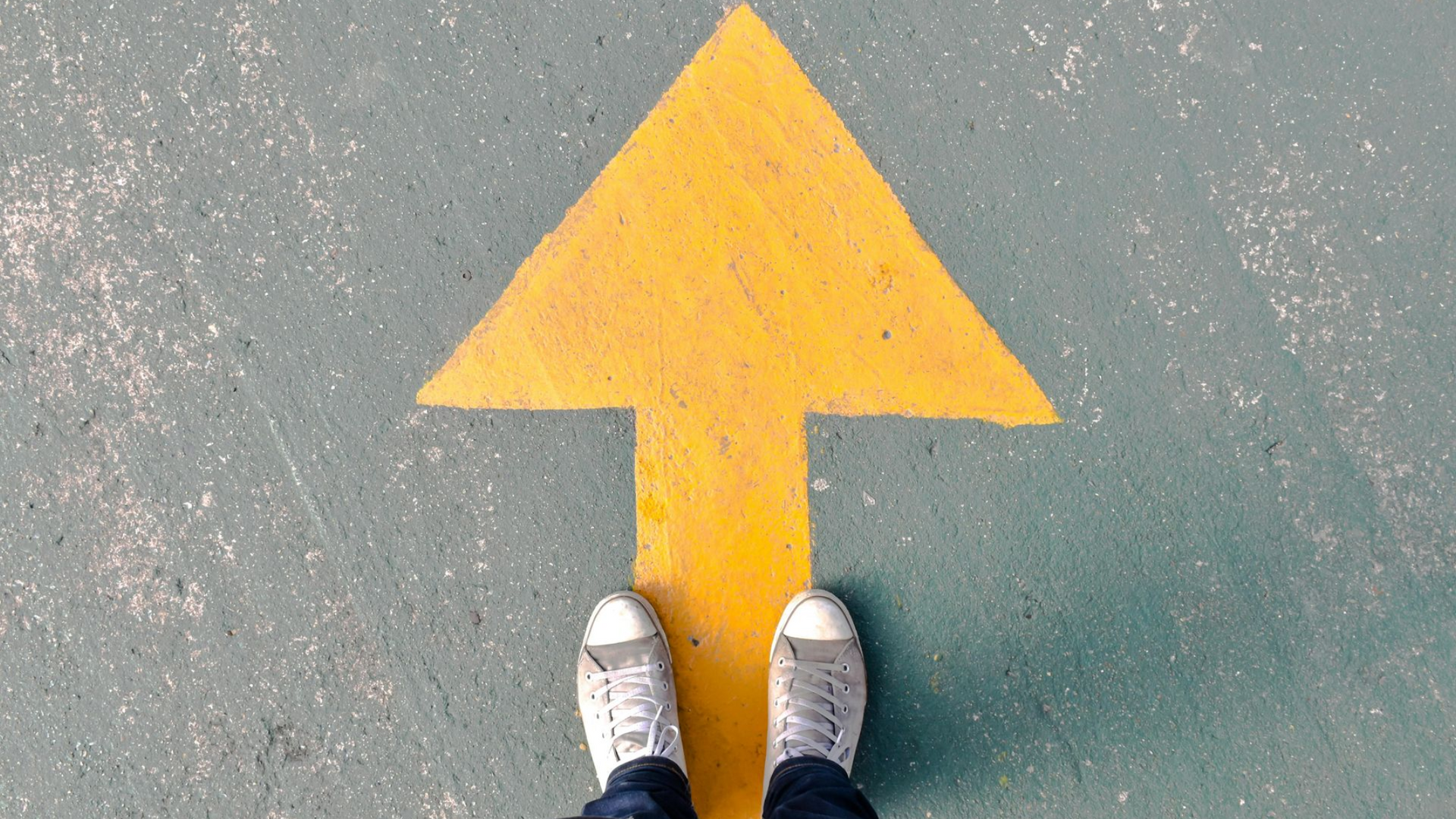 Feet in white sneakers standing on pavement beside a large yellow arrow pointing forward.