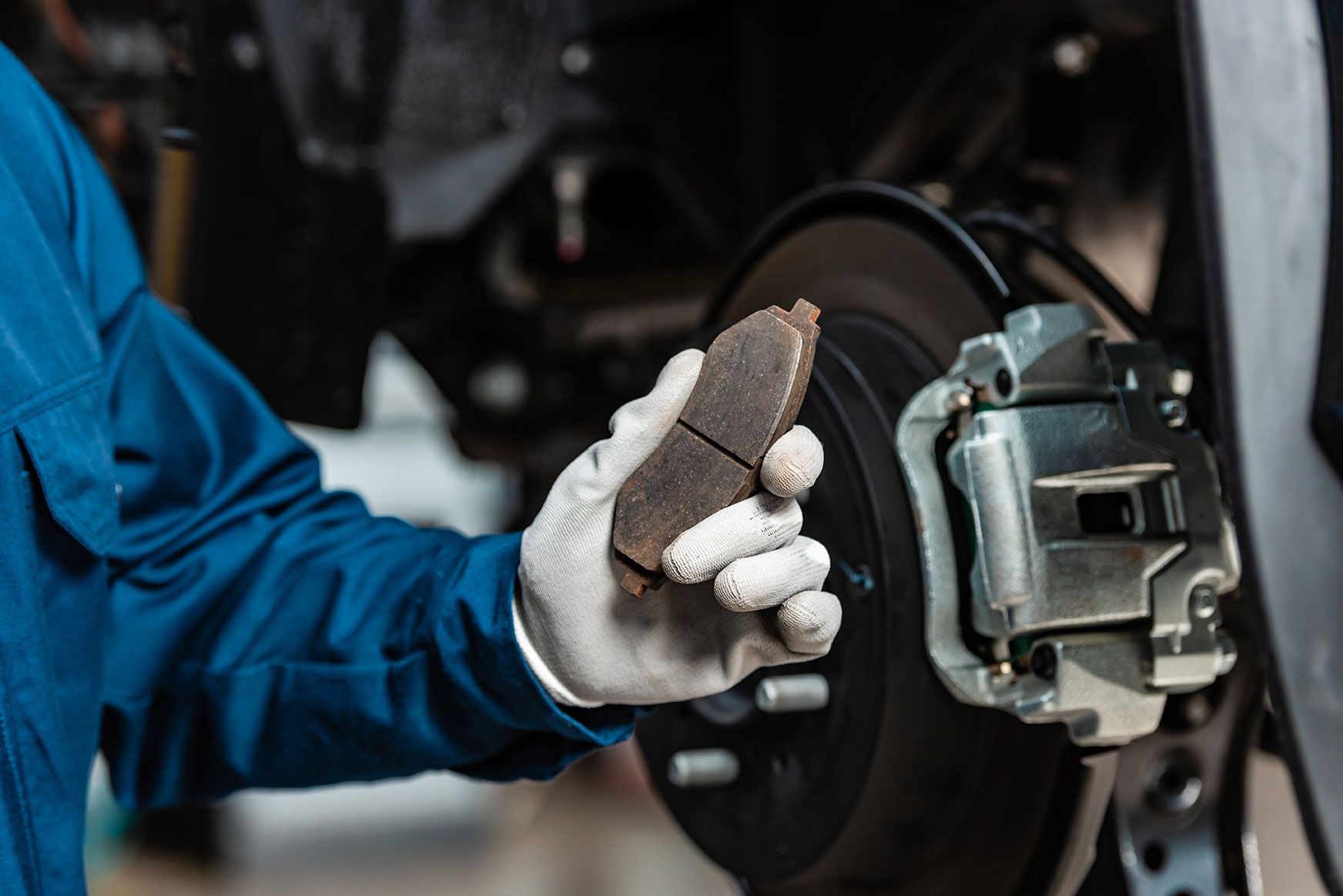 Mechanic in blue coveralls holding worn brake pad near a car's brake disc.