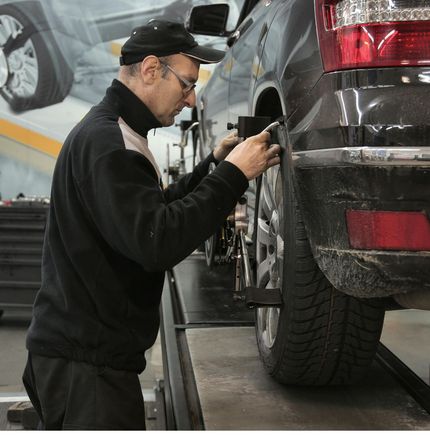 Mechanic in black uniform, cap, and glasses, checking car tire on a lift.