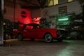 Red classic car in a dimly lit garage, bathed in red and green light, surrounded by car parts.