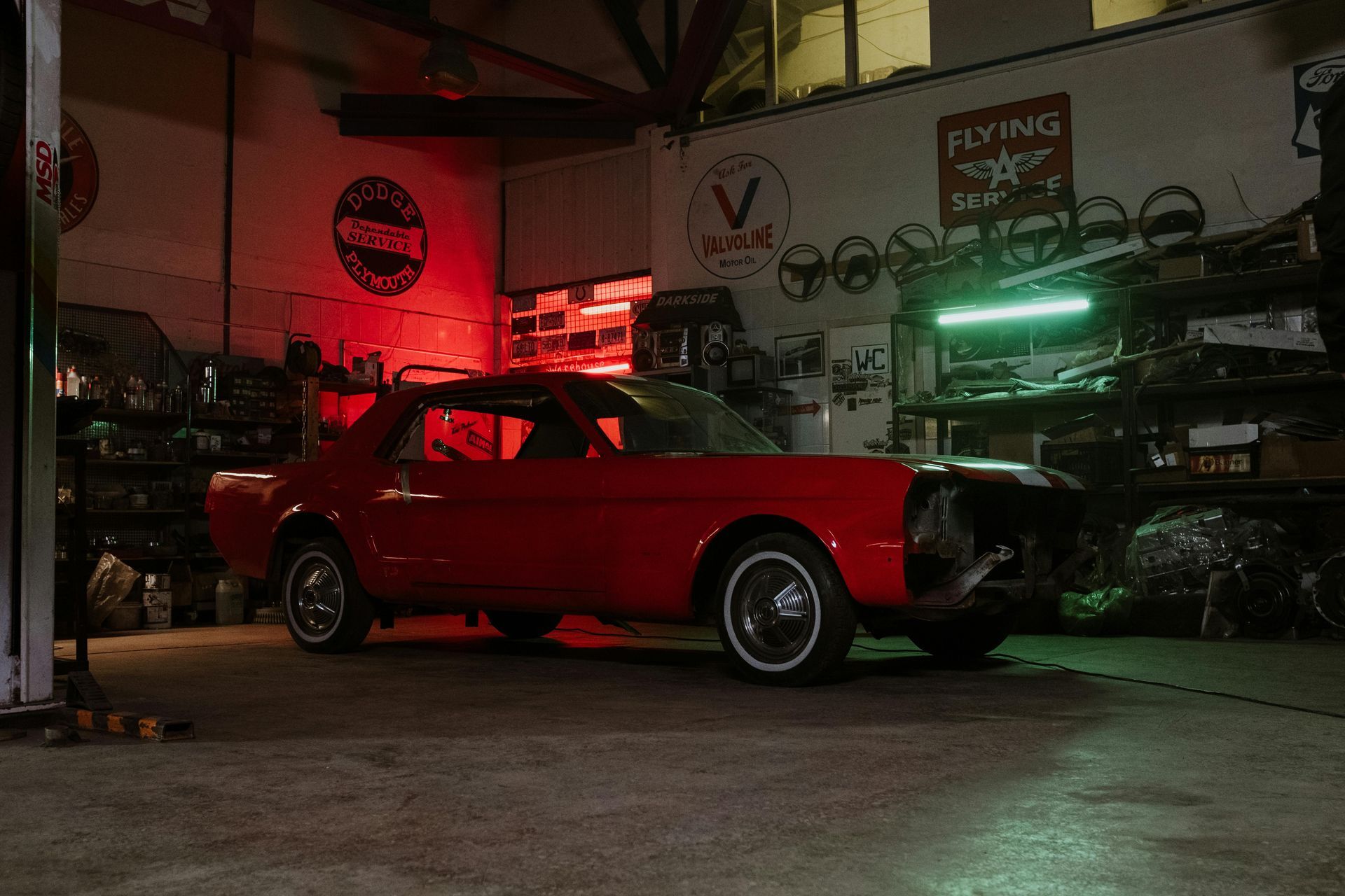 Red classic car in a dimly lit garage, bathed in red and green light, surrounded by car parts.