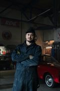 Mechanic in blue overalls, arms crossed, in a garage with a red classic car.