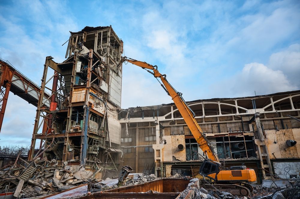 Excavator demolishing a weathered industrial building under a cloudy sky.