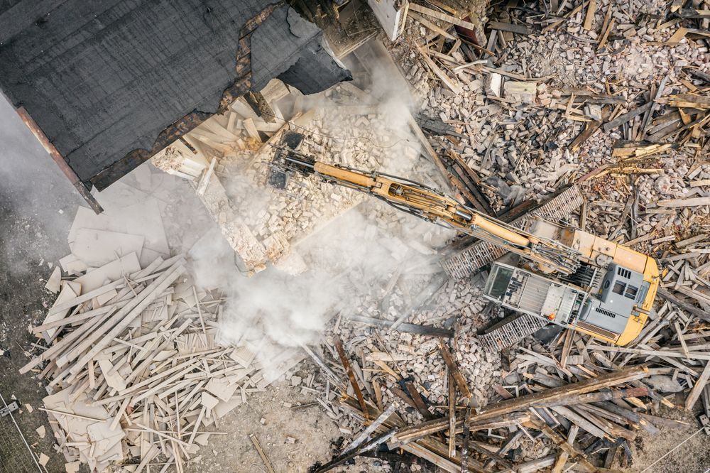 Overhead view of a yellow excavator demolishing a building, surrounded by debris and dust.