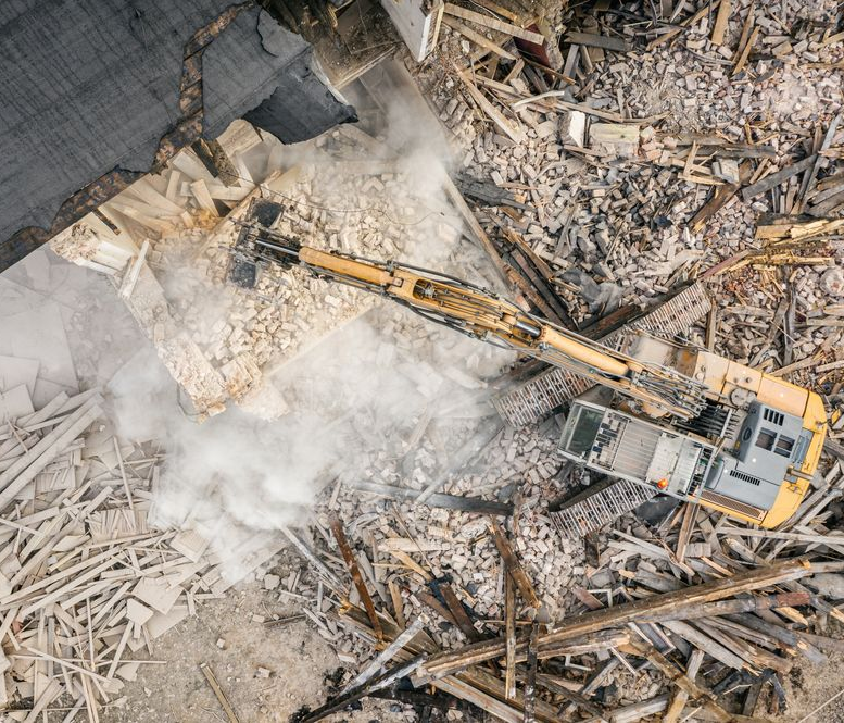 Overhead view of a yellow excavator demolishing a building, surrounded by debris and dust.