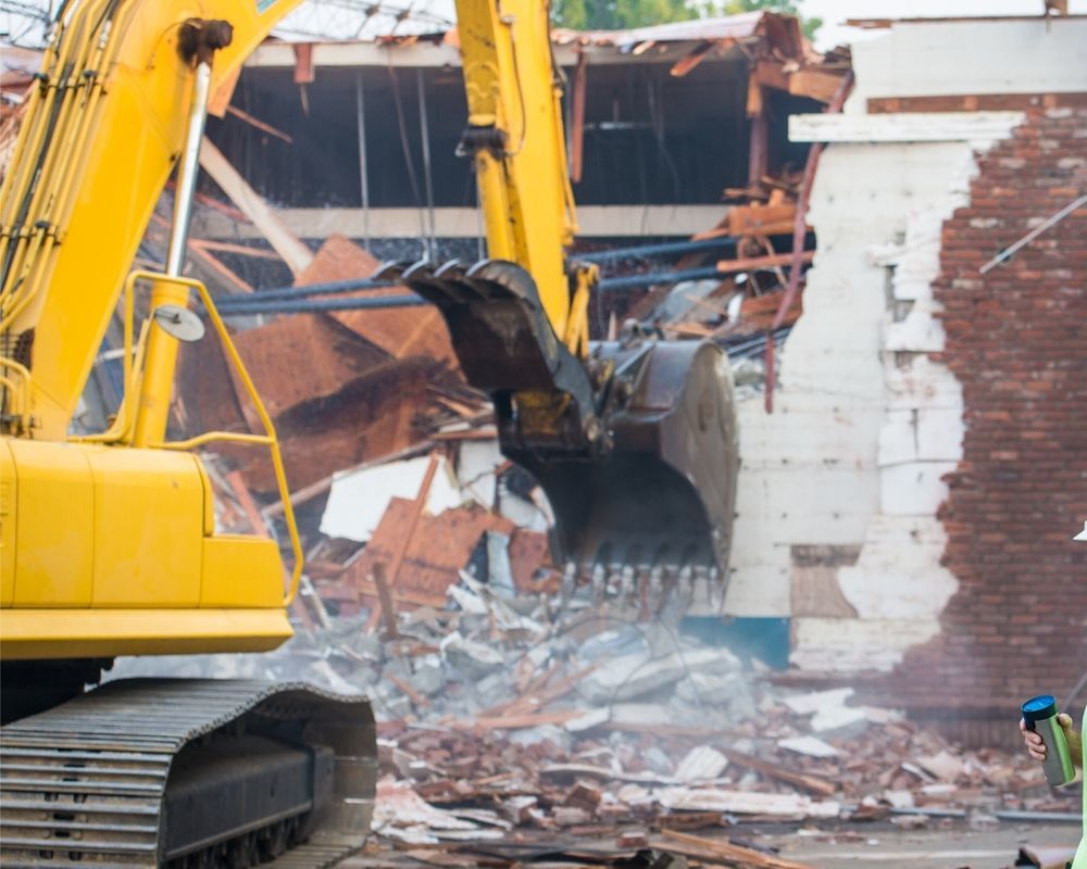 Controlled demolition of brick building with excavator removing structure and scattering debris during urban redevelopment