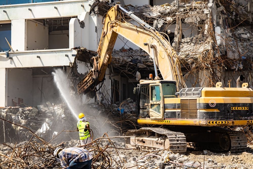 Dust suppression during demolition as excavator with hydraulic hammer breaks old building while worker sprays water to control dust