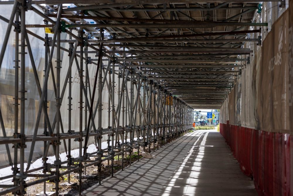 Overhead protection with covered pedestrian walkway at active construction site casting geometric shadows along sidewalk