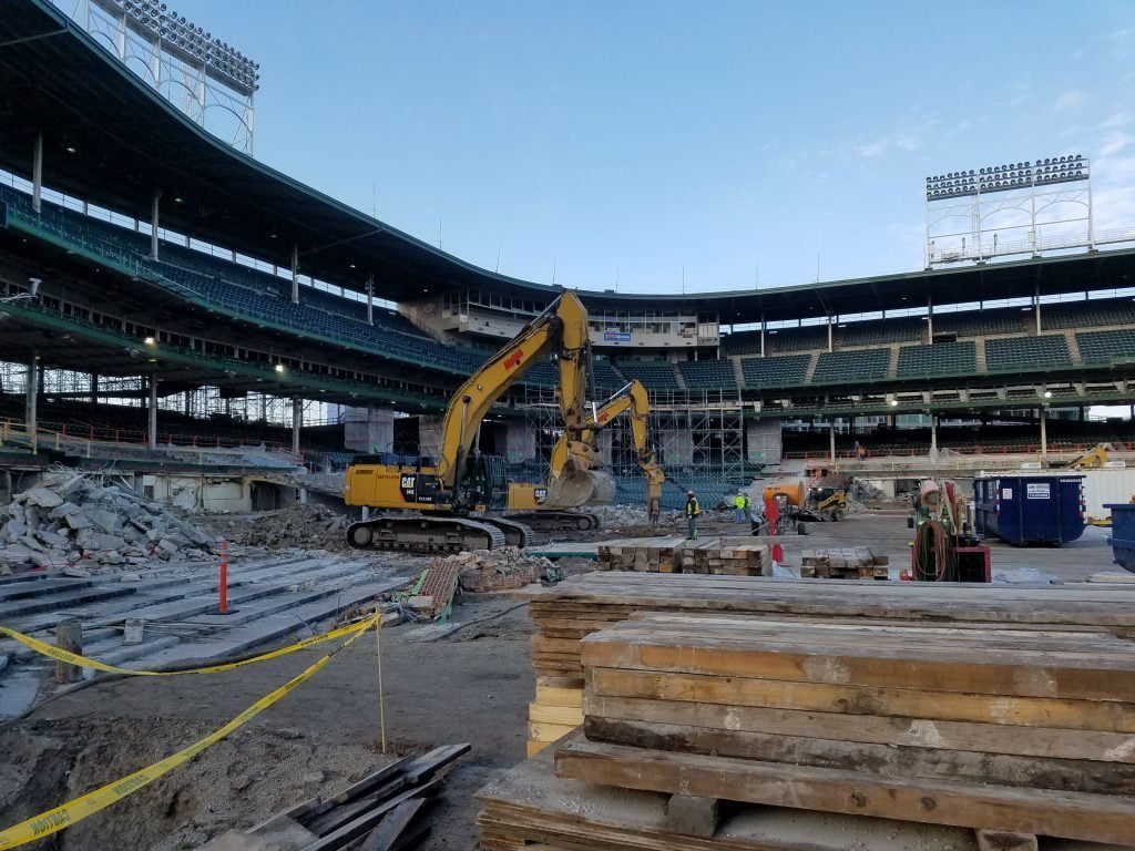 A large yellow excavator is digging in the dirt in front of a large stadium.