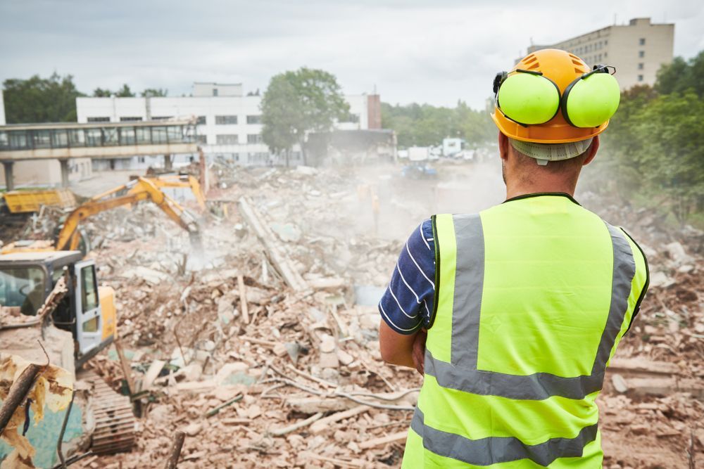 A large yellow excavator is digging in the dirt in front of a large stadium.