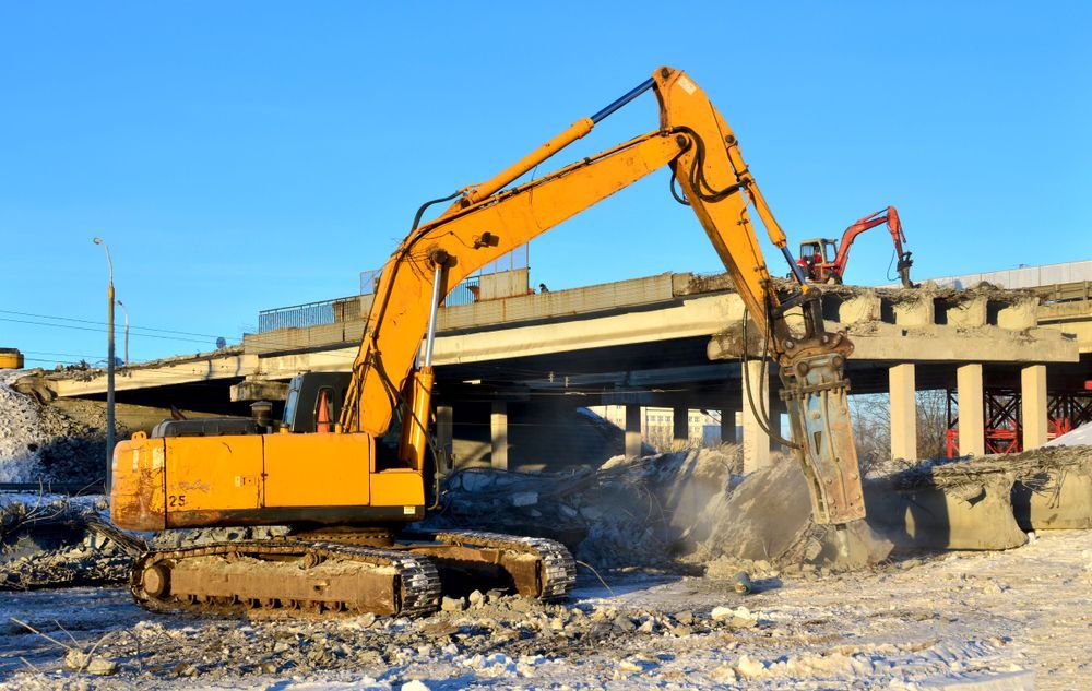 A yellow excavator is demolishing a bridge in the snow.