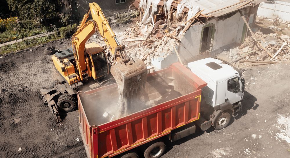 an excavator is demolishing a building and dumping the rubble into a dump truck .