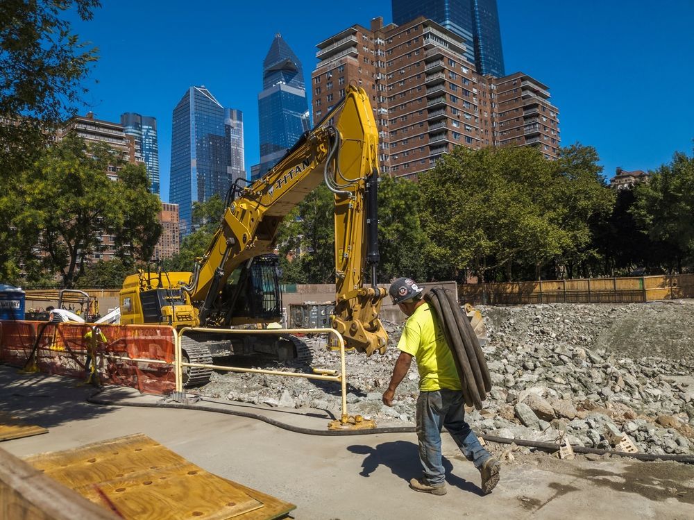 A construction worker is walking in front of a large yellow excavator.