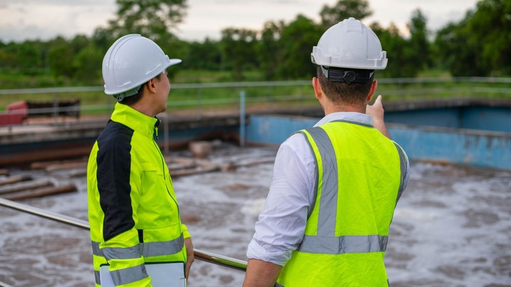 Two people in safety vests and hard hats at a water treatment facility; one points at a tank.