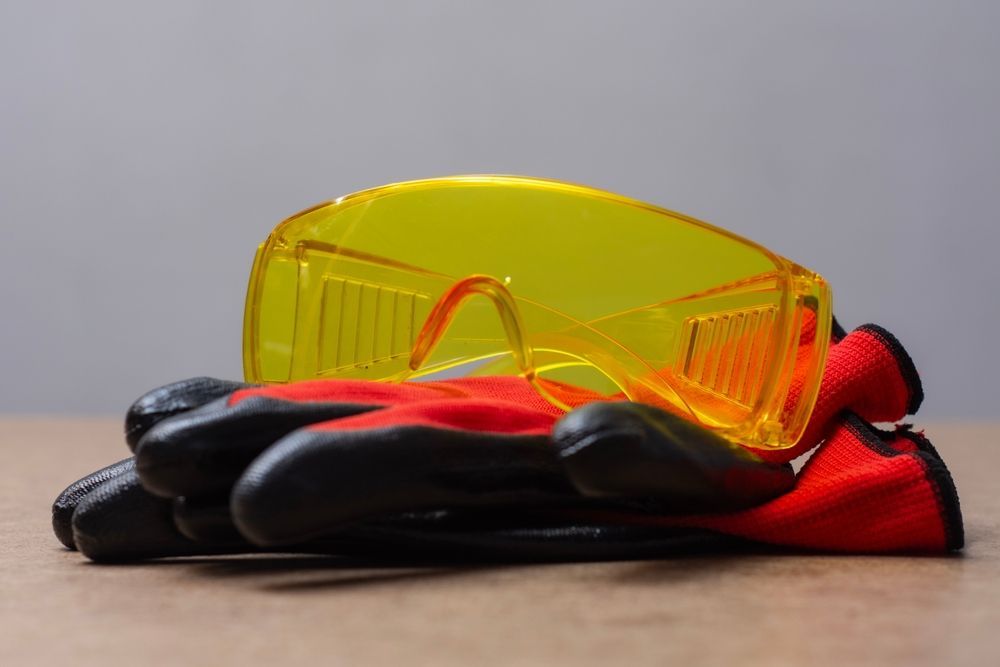 Yellow safety glasses atop red and black work gloves on a wooden surface.