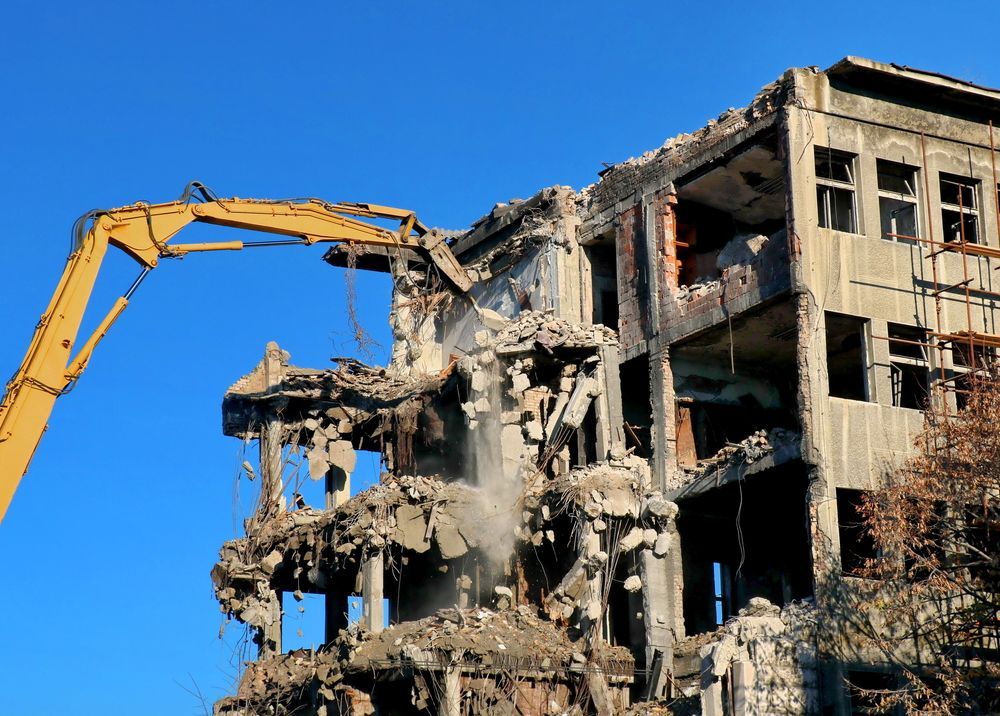 A large building is being demolished by a yellow excavator.