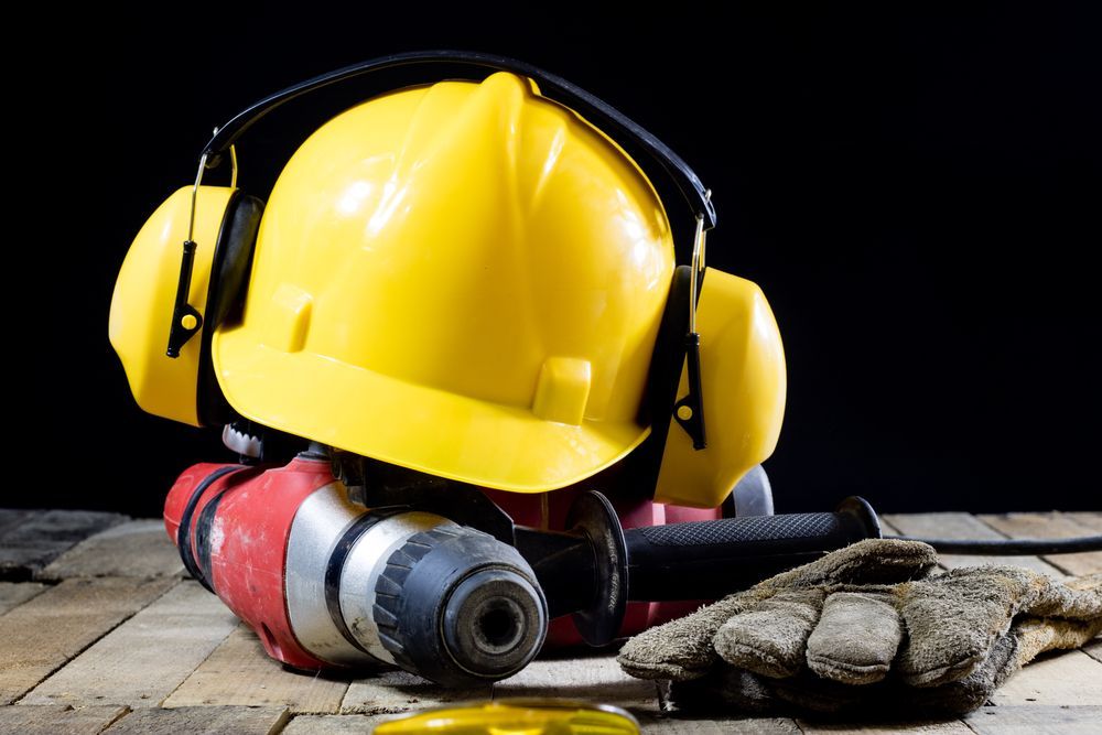 Yellow hard hat with earmuffs on a red drill and work glove.