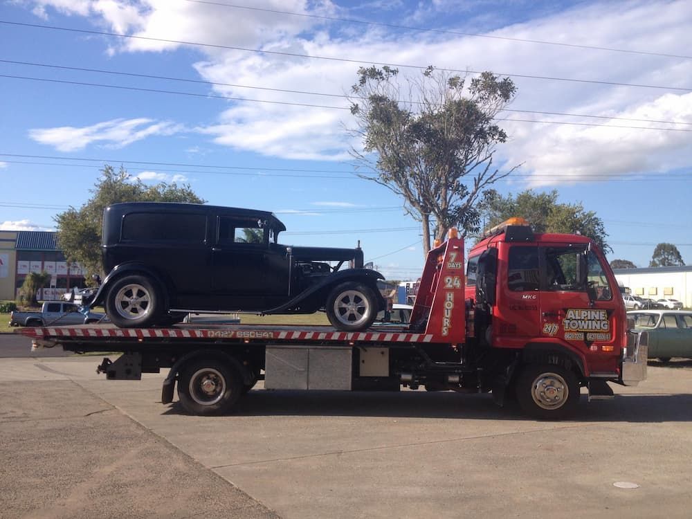 A Black Classic Car Is Being Towed by A Tow Truck — Alpine Towing Services in Lismore, NSW