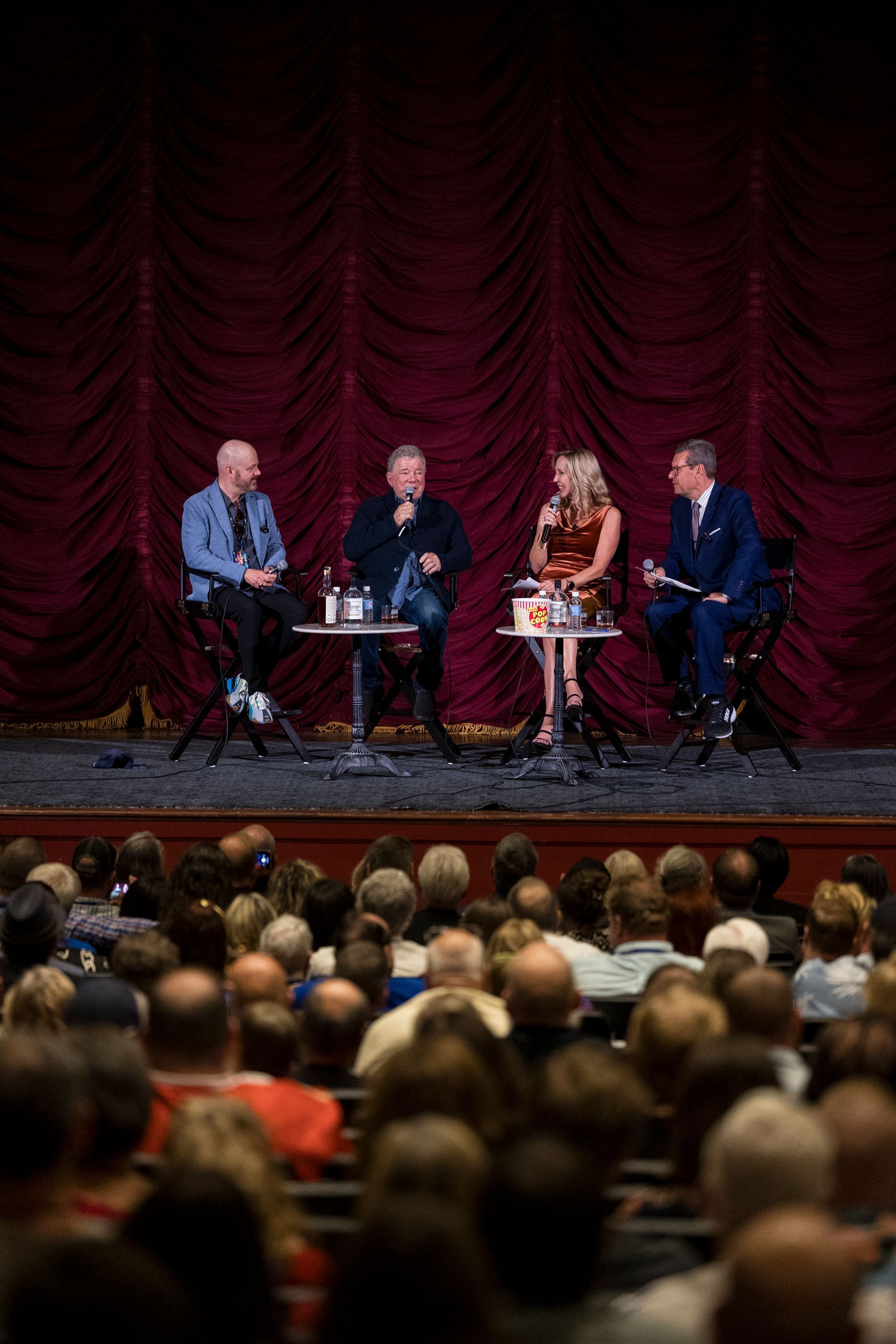 Four panelists seated on stage before a red curtain, speaking to an audience.