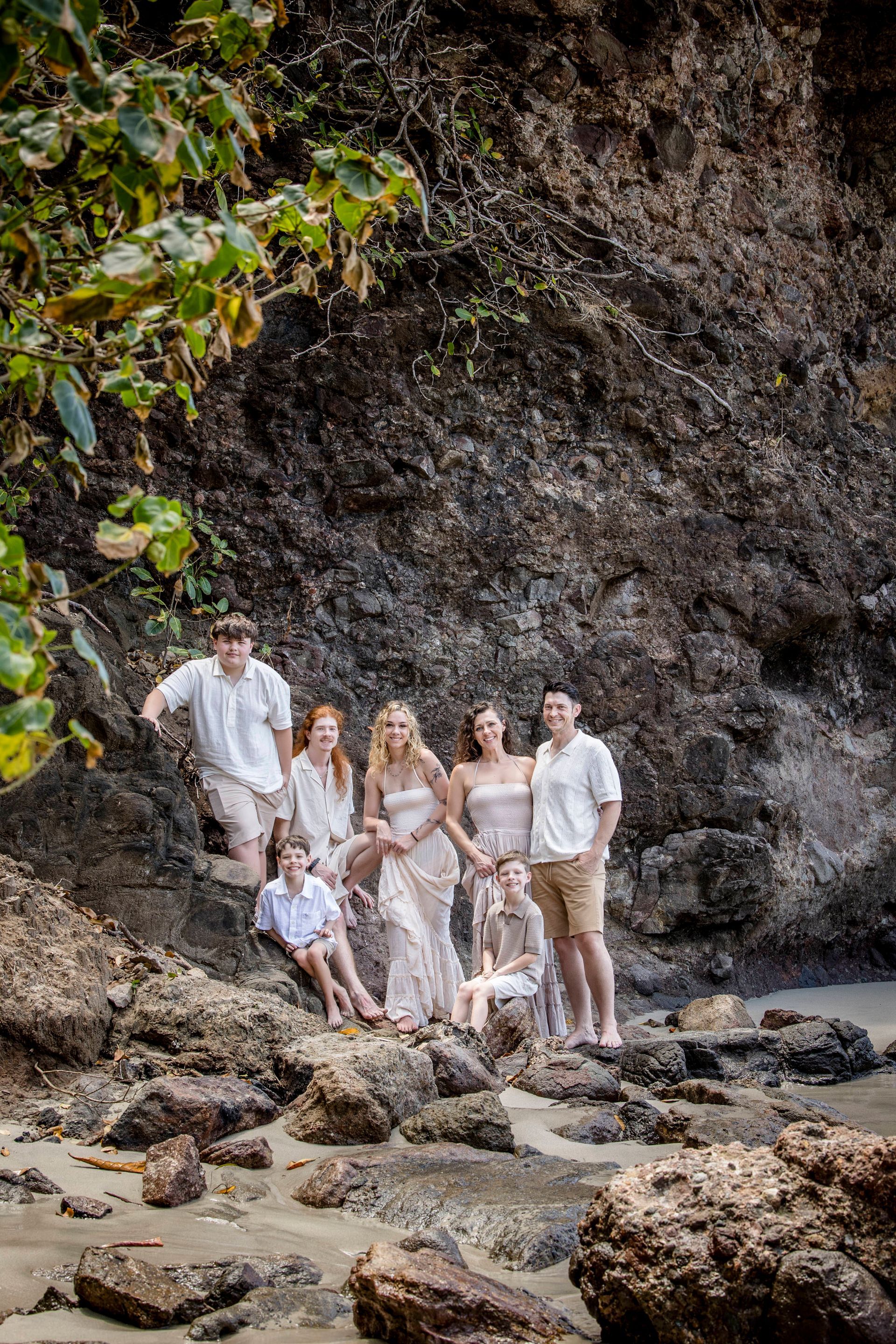 Group posing on a rocky beach beneath a cliff, surrounded by tropical greenery.