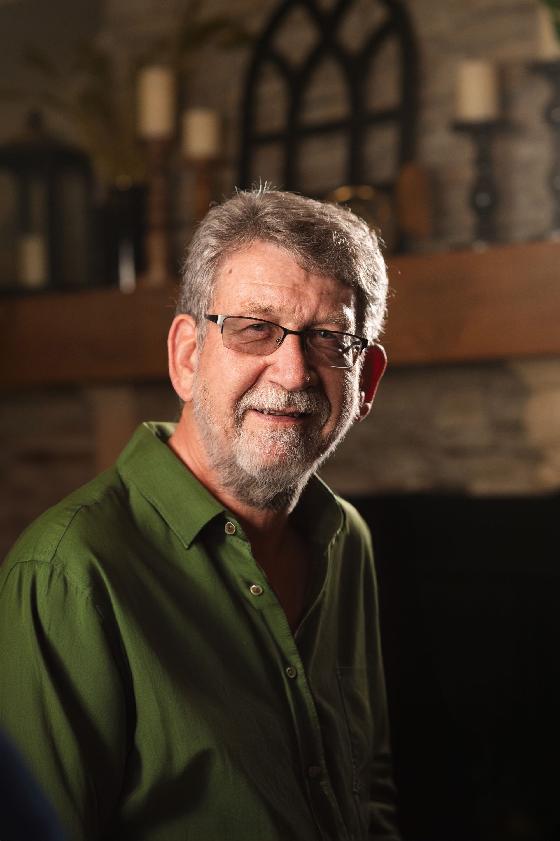 A man wearing glasses and a green shirt is standing in front of a fireplace.