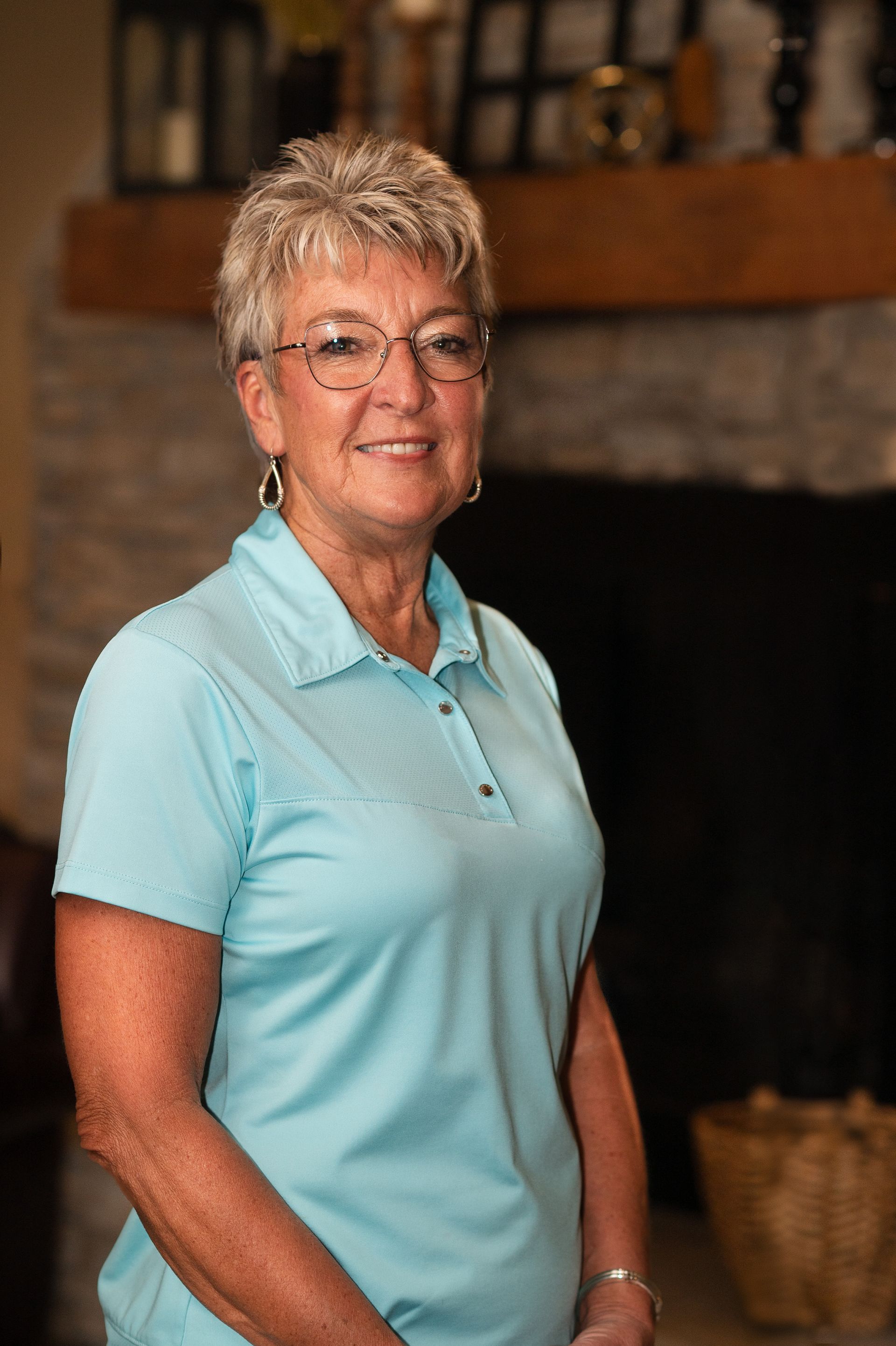 A woman in a blue shirt and glasses is standing in front of a fireplace.