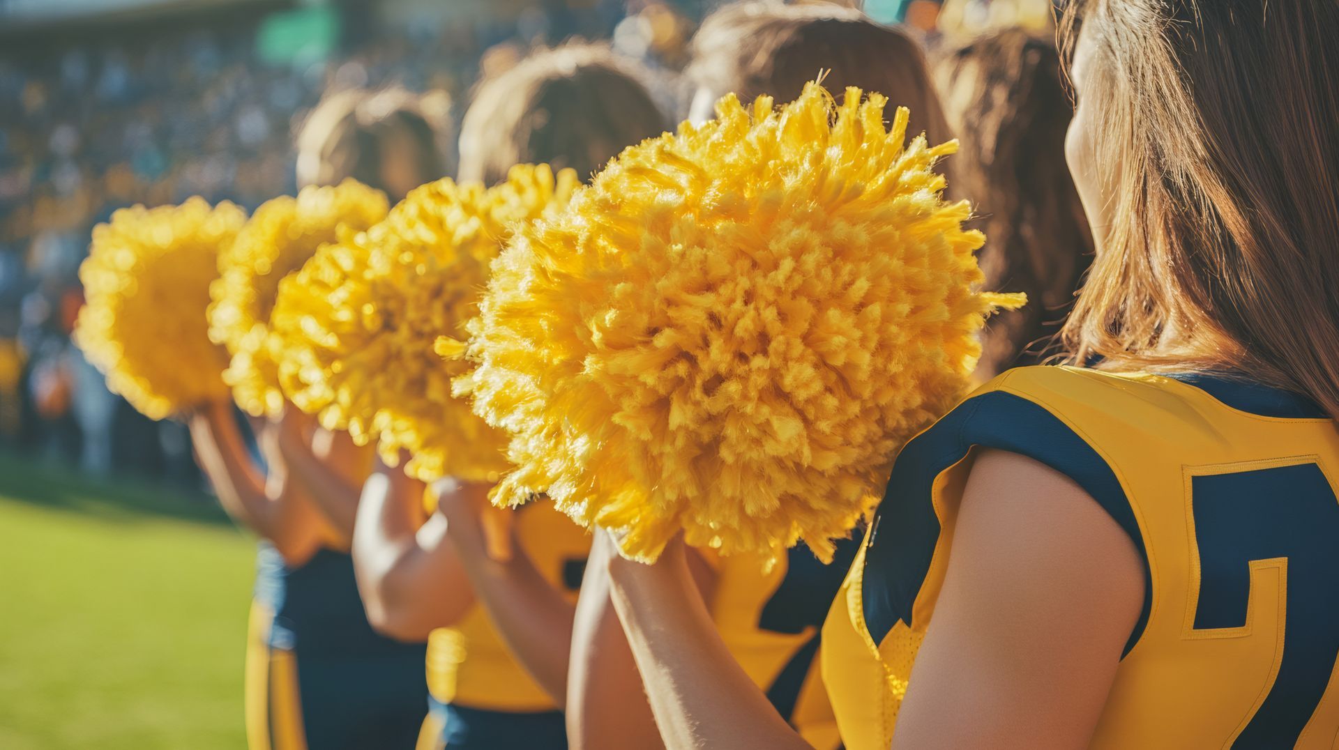 Cheerleaders in gold and blue uniforms hold yellow pom-poms on a sunny sports field.