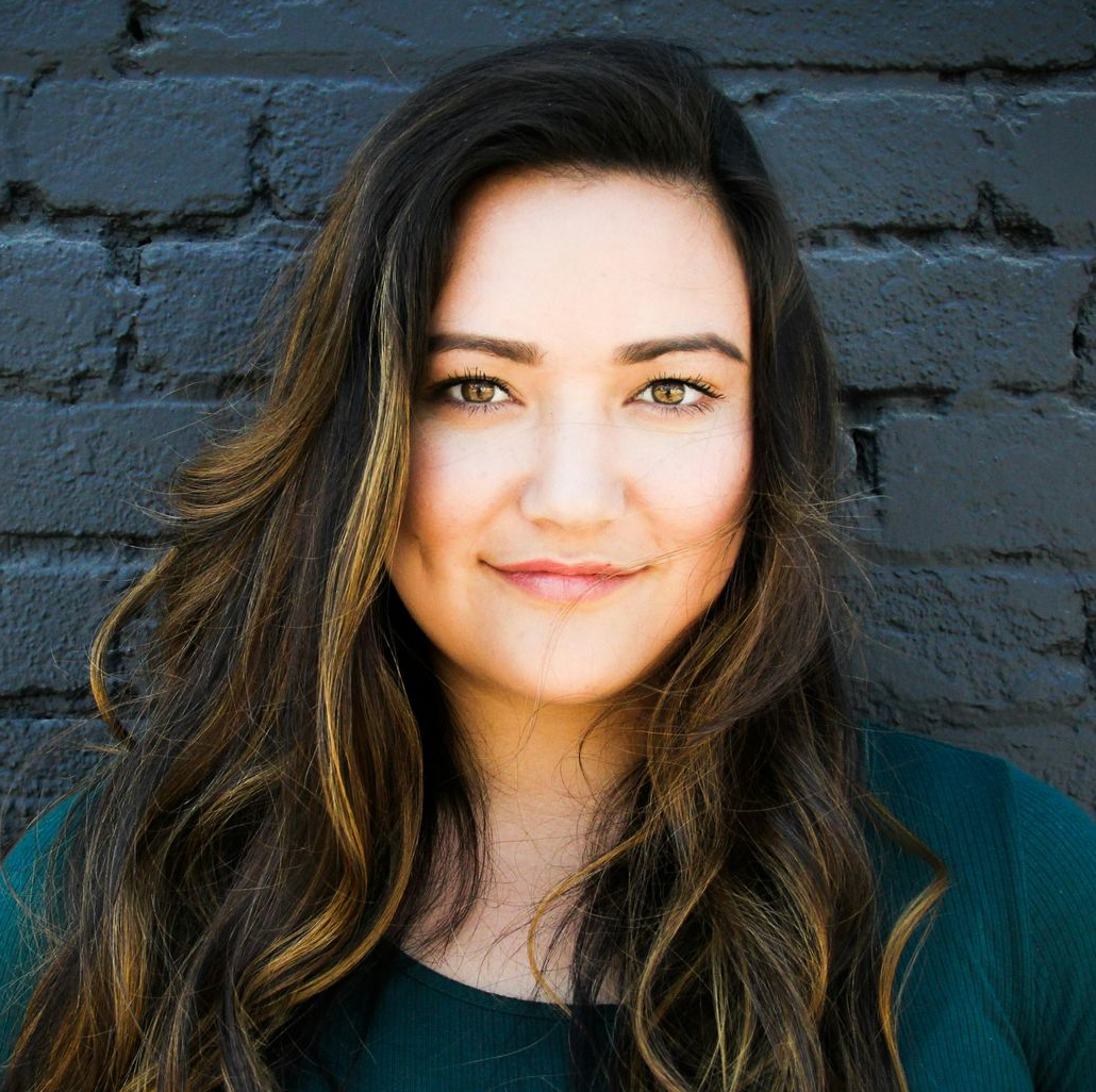 Woman with long, wavy brown hair, smiling, against a dark brick wall.