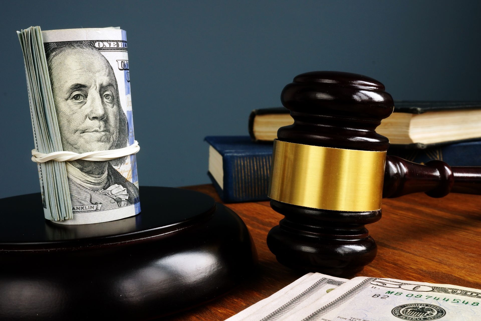 Photograph showing a wooden judge's gavel with a gold band resting on a table alongside a stack of law books and a roll of U.S. dollar bills secured with a rubber band