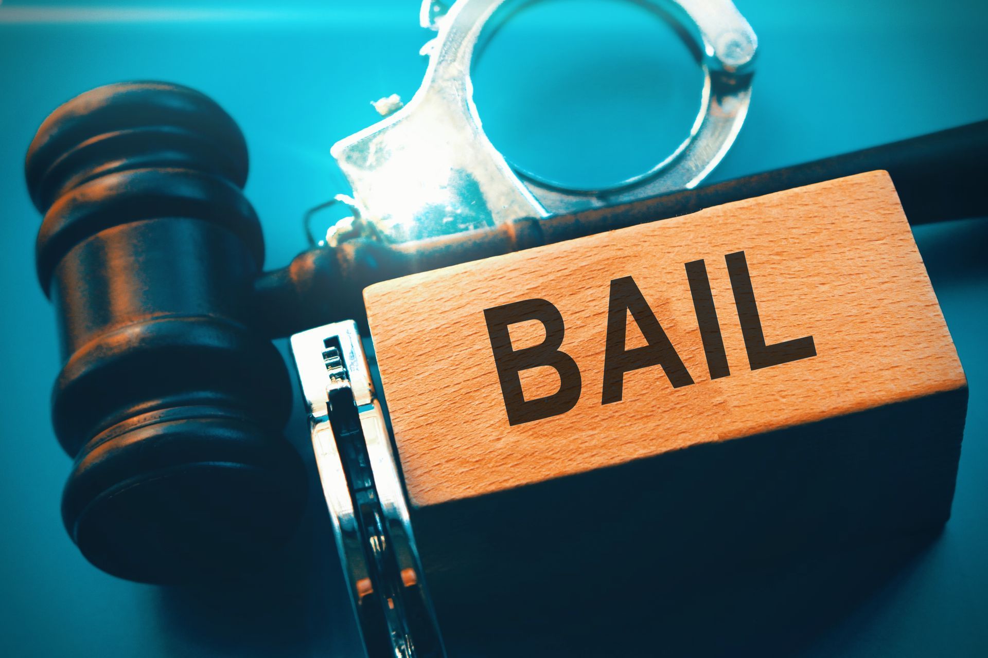 Wooden block labeled ‘Bail’ placed beside a gavel and metal handcuffs on a blue surface.
