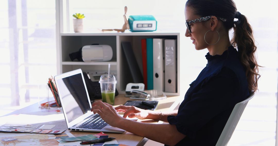 Woman typing on laptop at desk, wearing glasses, in front of a shelf with items.