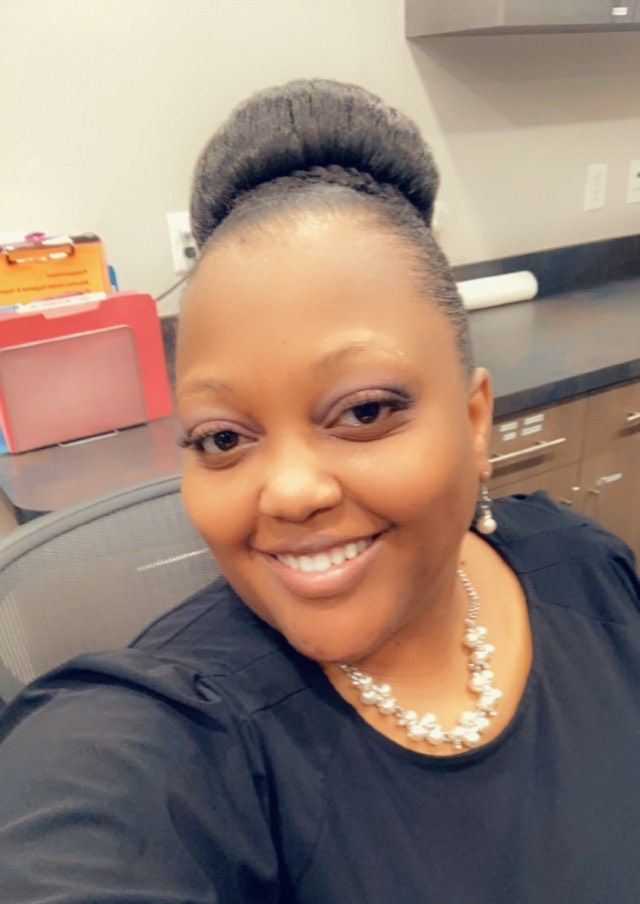Woman with bun smiling, wearing pearl necklace and black top, indoors.