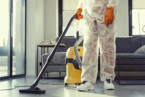 Person in protective suit vacuums a room, yellow vacuum cleaner, orange gloves, white sneakers.