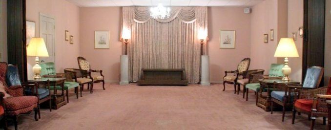 A symmetrical, formal waiting room with light pink walls, plush carpeting, armchairs, lamps, and draped windows.