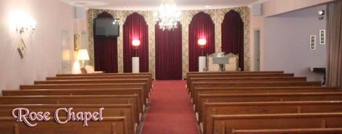 A formal chapel with rows of wooden pews facing a stage decorated with red curtains, white pedestals, and a chandelier.