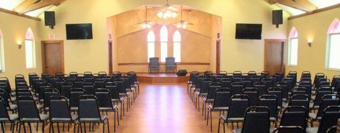 An indoor meeting space with rows of black chairs facing a wooden stage with two chairs in front of three arched windows.