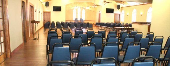 A large, empty event hall with rows of blue chairs facing a stage with two chairs in front of arched windows.