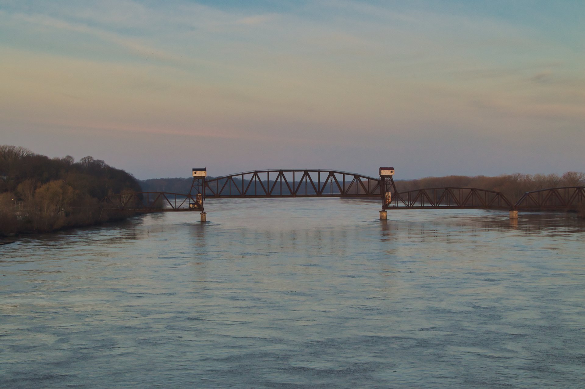 A dark metal railway bridge spans a wide, flowing river under a soft, dusk-lit sky.