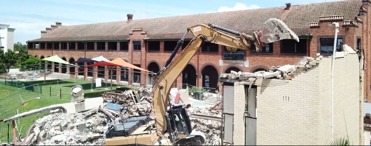 A large brick building is being demolished by an excavator