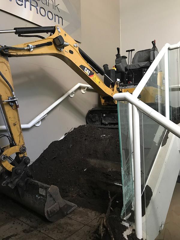A small excavator is digging a hole in the floor next to stairs.