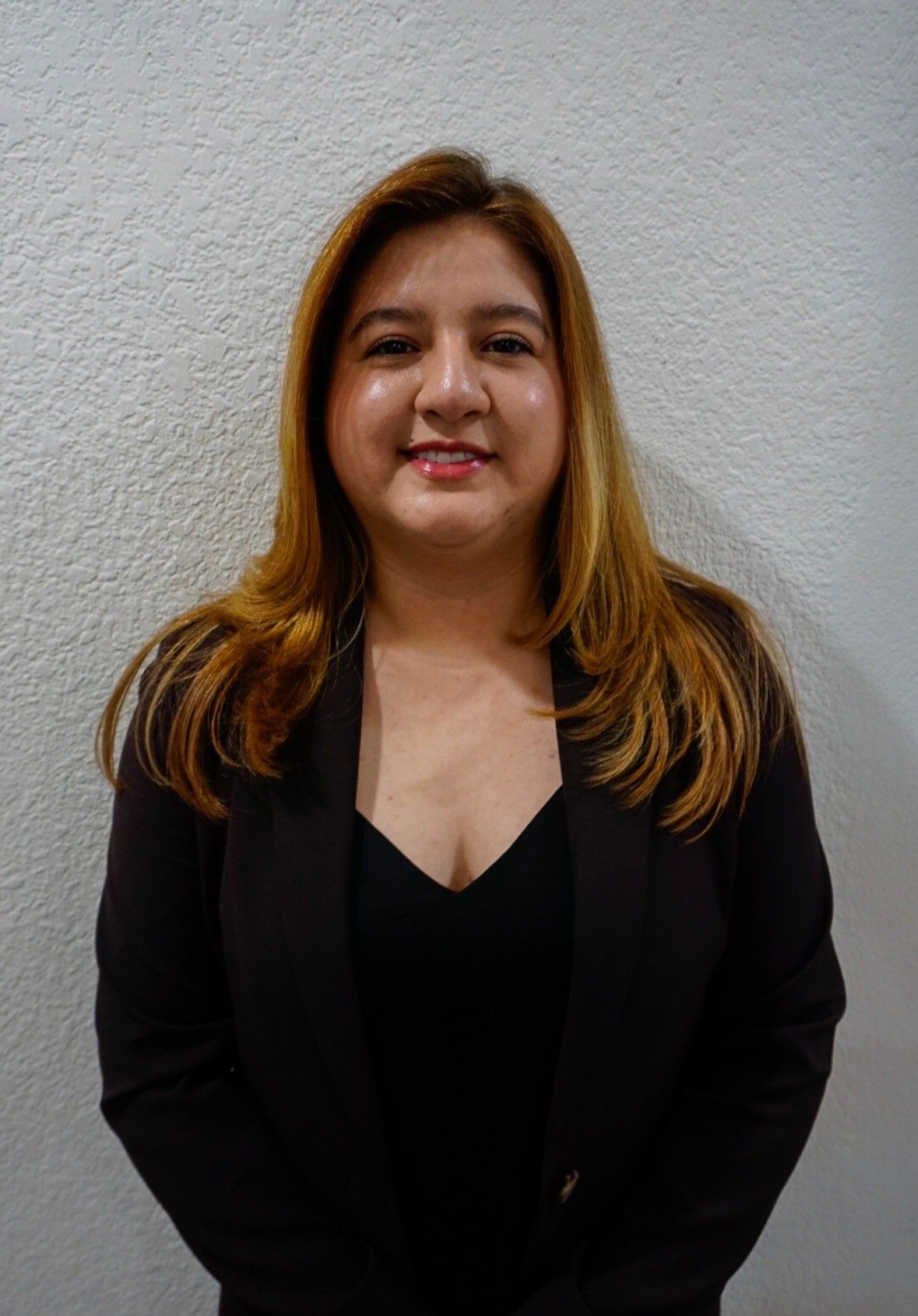 Woman with brown hair in a dark suit, smiling, standing in front of a white wall.